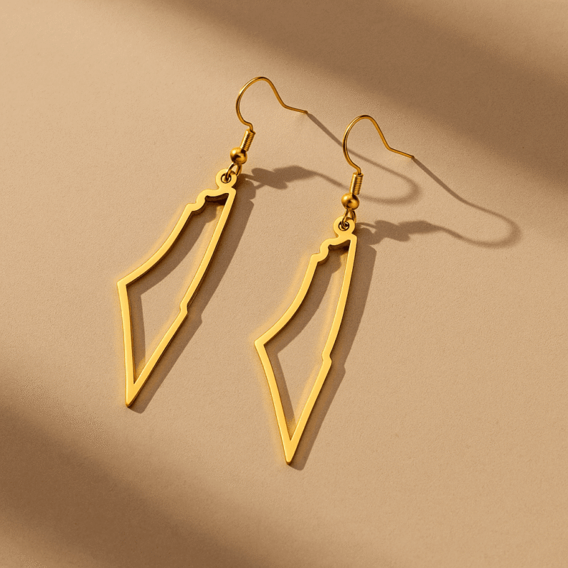 Deux boucles d’oreilles dorées en forme de Palestine posées à plat sur fond beige